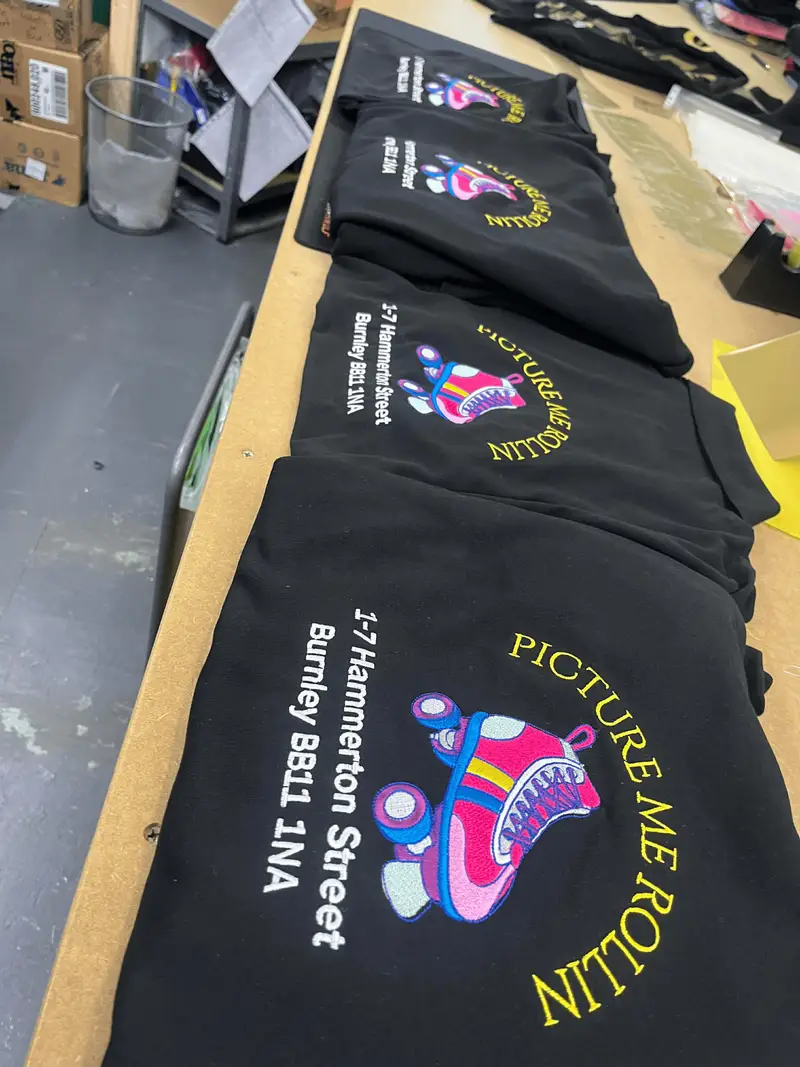 A run of black hoodies on the workbench, each embroidered with the Picture Me Rollin roller skate logo and Burnley address