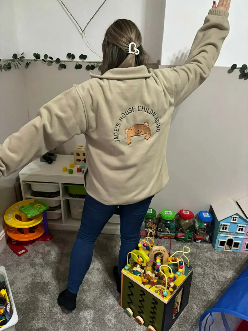 Beige fleece embroidered on the back with the Jade's House Childminding bear logo, worn in a playroom