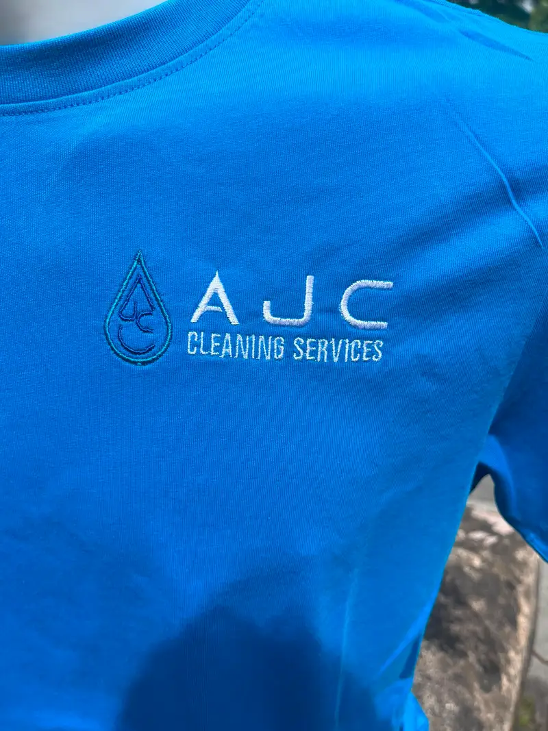 Close-up of the AJC Cleaning Services droplet logo embroidered in white on a blue t-shirt