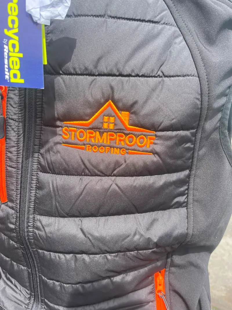Close-up of the Stormproof Roofing logo embroidered in orange thread on a black padded gilet at the ThreeKZ workshop in Colne