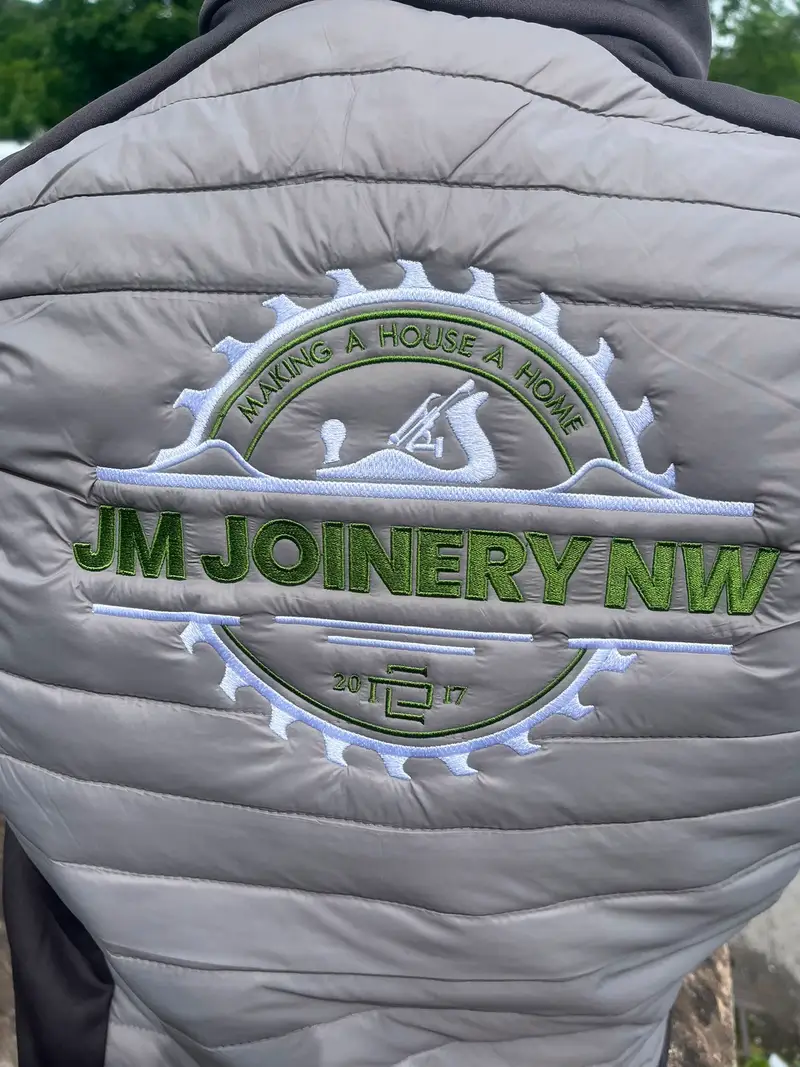 Large JM Joinery NW cog logo embroidered in green and white across the back of a grey gilet at the ThreeKZ workshop in Colne