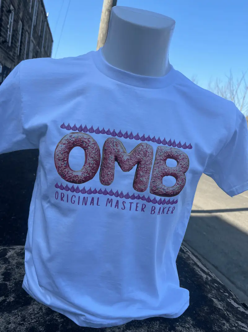 White t-shirt printed with the OMB Original Master Baker logo, the letters styled as doughnuts